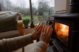 Hands warming near a wood-burning stove in a cosy British living room with a rainy window in the background