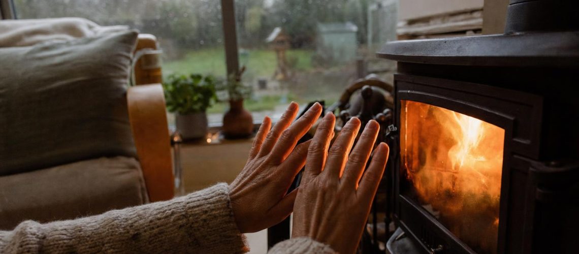 Hands warming near a wood-burning stove in a cosy British living room with a rainy window in the background
